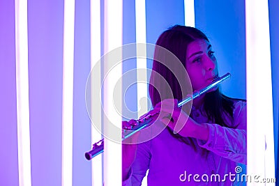 Young pretty woman plays the flute. Musician female posing in a dark studio against the backdrop of bright neon lights Stock Photo