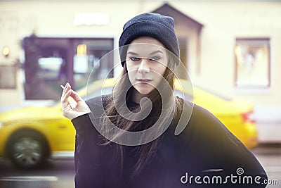 Young pretty woman smoking outside. Hipster outfit, wearing black hat and t-shirt, city yellow taxi on the background. Stock Photo