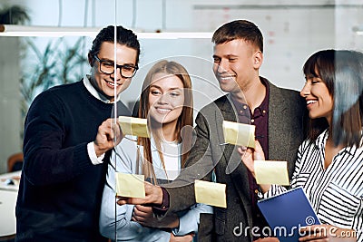 Young professional team. Two men and two women gluing stickers on glass wall in creative office. Coworking and Stock Photo