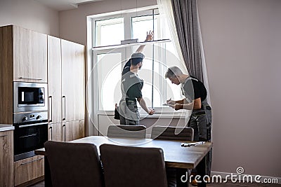 Young professional worker in uniform using tape measure, measuring window for installing blinds, while his aged Stock Photo