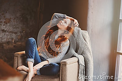 Young readhead woman relaxing at home in cozy chair, dressed in casual sweater and jeans Stock Photo