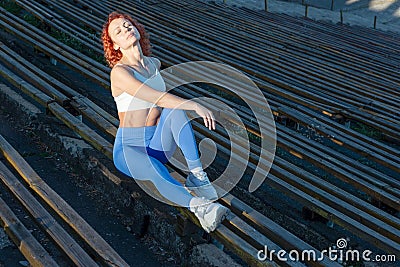 Young red-haired sportswoman sits on a wooden bench of a sports stadium in the rays of the setting sun. on a dark Stock Photo