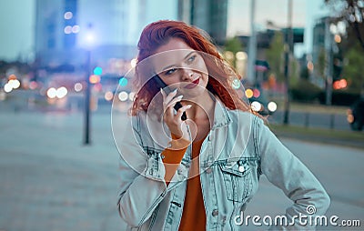 Young redhaired woman standing at night with phone by european skyline Stock Photo