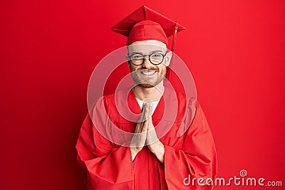 Young redhead man wearing red graduation cap and ceremony robe praying with hands together asking for forgiveness smiling Stock Photo