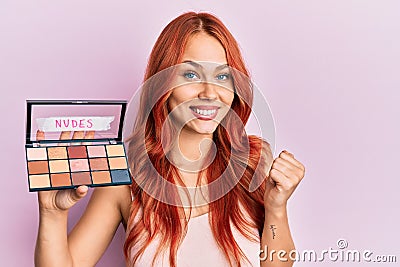 Young redhead woman holding makeup nudes screaming proud, celebrating victory and success very excited with raised arm Stock Photo