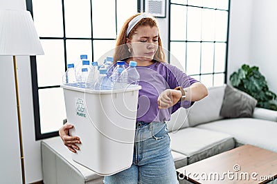 Young redhead woman holding recycling wastebasket with plastic bottles checking the time on wrist watch, relaxed and confident Stock Photo