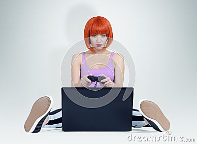 Young redhead woman with joystick sits on the floor in front of a laptop. Gamer plays Stock Photo