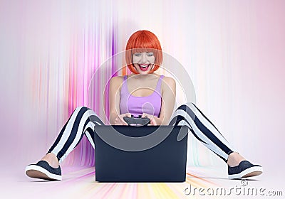 Young redhead woman with joystick sits on the multicolored floor in front of a laptop. Gamer plays. Stock Photo