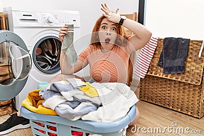 Young redhead woman putting dirty laundry into washing machine surprised with hand on head for mistake, remember error Stock Photo