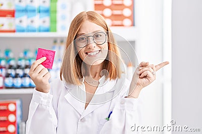 Young redhead woman working at pharmacy drugstore holding condom smiling happy pointing with hand and finger to the side Stock Photo