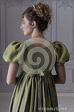 A young Regency woman wearing a green shot silk dress and standing in front of a white paneled wall shown in back view Stock Photo