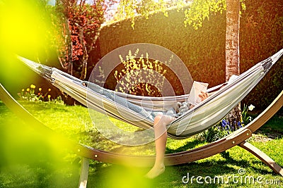 Young relaxed girl reading book in hammock in garden at home at bright sunset. Slow living, Stock Photo