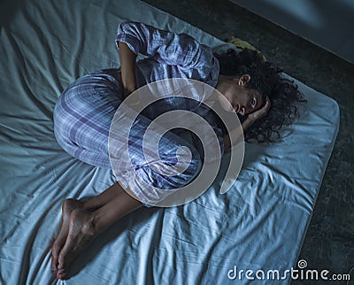 Young sad and depressed hispanic woman with curly hair sleepless in bed suffering excruciating period pain holding her belly in Stock Photo