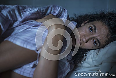 Young sad and depressed hispanic woman with curly hair sleepless in bed suffering excruciating period pain holding her belly in Stock Photo