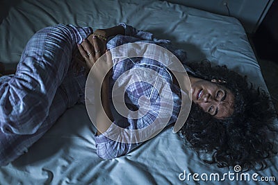 Young sad and depressed latin woman with curly hair sleepless in bed suffering excruciating period pain holding her belly in fetal Stock Photo