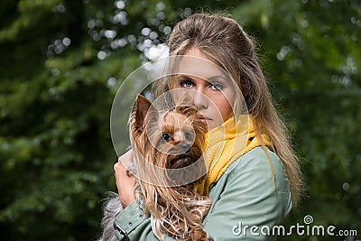 Young sad pretty blonde woman in city park. Small yorkshire terrier is on her hands. Stock Photo