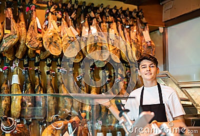 Young salesman in black apron offering dry-cured jamon in butcher shop Stock Photo