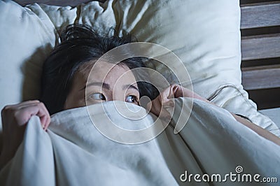 Young scared and stressed Asian Chinese woman lying in bed suffering nightmare in fear and panic grasping blanket covering her hor Stock Photo
