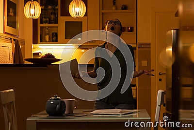 Young screaming shocked Caucasian woman wearing glasses holding documents while standing in kitchen. Concept of overdue Stock Photo