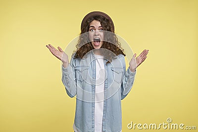 Young screaming and surprised brunette woman with curly hair and hat, happy about new present, isolated over yellow Stock Photo