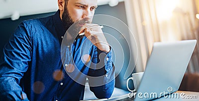 Young bearded businessman sitting in front of computer, looking at screen, holding pen, thinking. Freelancer works home. Stock Photo