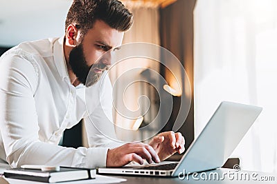 Young serious bearded businessman standing in office near table and using laptop. Man works on computer, checks e-mail Stock Photo