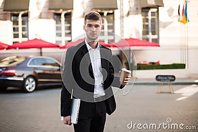 Young serious man in black suit and white shirt with wireless earphones holding laptop and coffe in hands while Stock Photo