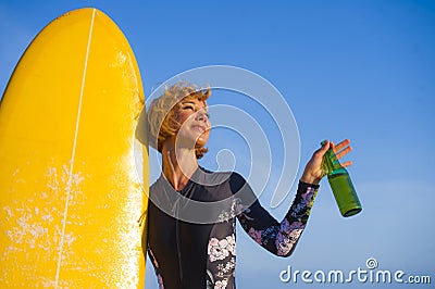 Young beautiful and happy surfer girl holding yellow surf board smiling cheerful drinking beer bottle enjoying summer holiday Stock Photo