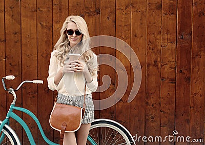 Young blonde girl with long hair with brown vintage bag in sunglasses standing near vintage green bicycle and holding a cup o Stock Photo