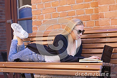 Young sexy girl with blond hair in sunglasses works on a laptop while lying on a bench near a brick wall of a building Stock Photo