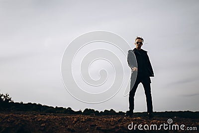 Young sexy man in leather jacket and sunglasses standing outdoor. Dark silhouette against grey sky Stock Photo