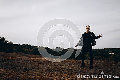 Young sexy man in leather jacket and sunglasses standing outdoor. Dark silhouette against grey sky Stock Photo