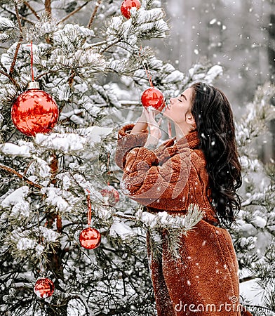Young sexy brunette woman in stylish fur coat stands near decorated Christmas tree kisses red balls under snow Stock Photo