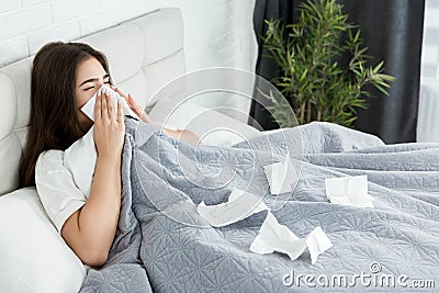 Young sick woman sitting in bed bowing out her running nose with paper towel feeling bad early in the morning Stock Photo