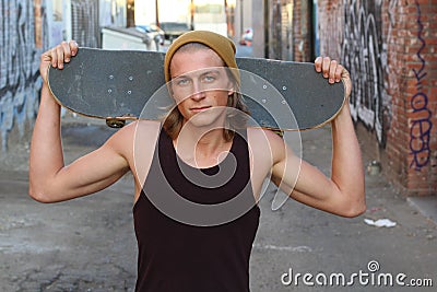 Young skater in a tank top shirt and beanie holding a skateboard over his shoulder and looking at the camera isolated on alleyway Stock Photo