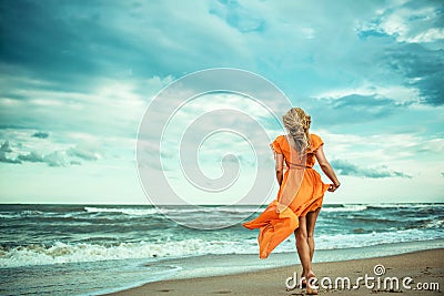 A young slender woman in orange dress is walking barefoot towards the storming sea Stock Photo