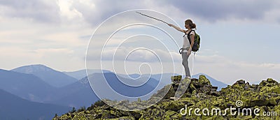 Young slim blond tourist girl with backpack points with stick at foggy mountain range panorama standing on rocky top on bright blu Stock Photo