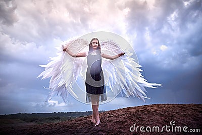 Young slim girl in black dress with with white wings dances on sand dunes against a dramatic sky before a thunderstorm Stock Photo