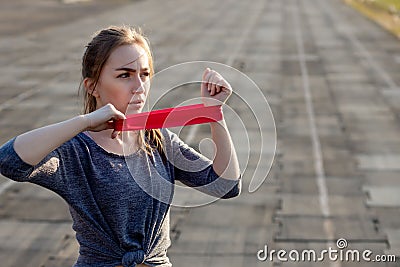 Young slim woman in sportswear doing squats exercise with rubber band on a black coated stadium track Stock Photo