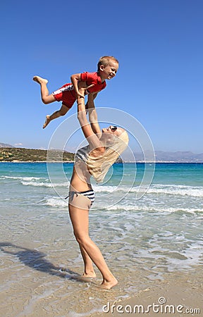 Young slim woman throws up and catches her little son on the beach by the clear blue sea. Mother and her cute little baby-boy are Stock Photo