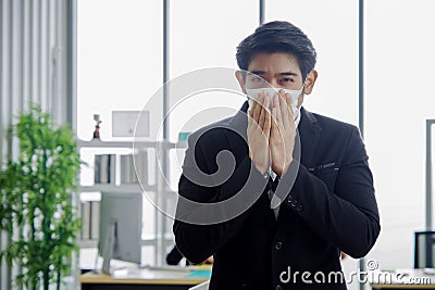 Young smart Asian businessman is sneeze and wearing a mask at workplace to prevent the corona virus outbreak Stock Photo