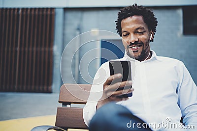 Young smiling african man using smartphone to listen to music while sitting on the bench at sunny street.Concept of Stock Photo