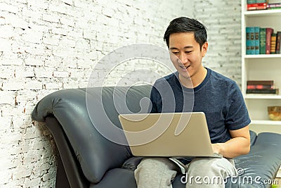 Young smiling Asian man in casual clothing on sofa working on laptop computer. Male freelancer working from home on Stock Photo