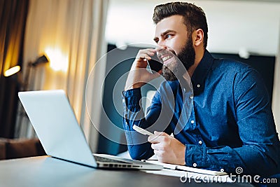 Young smiling bearded businessman sitting in front of computer, talking on cell phone, holding pen. Phone conversations. Stock Photo