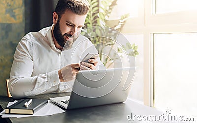 Young smiling bearded businessman sitting at table in front of computer, using smartphone. Man checking email, chatting. Stock Photo