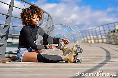 Young black girl sitting on urban bridge and puts on skates. Woman with afro hairstyle rollerblading on sunny day Stock Photo