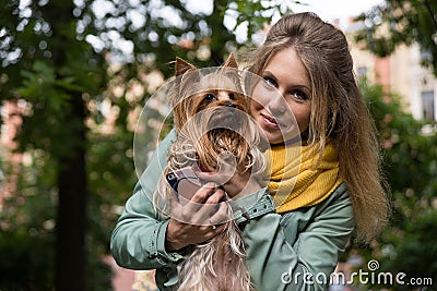 Young smiling blonde woman in city park. Small yorkshire terrier is on her hands. Stock Photo