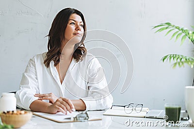 Young smiling brunette woman nutritionist plus size in white shirt working at laptop on table with house plant in bright modern Stock Photo