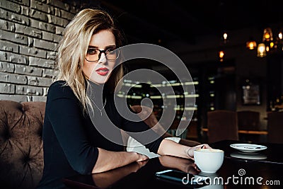 Young smiling business woman sitting in cafe at table, leaning hand on table and holding smartphone. Stock Photo