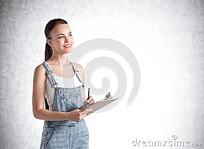 smiling girl in denim overalls holding a planner and dreaming about to do list of upcoming week. Concrete wall on background Stock Photo